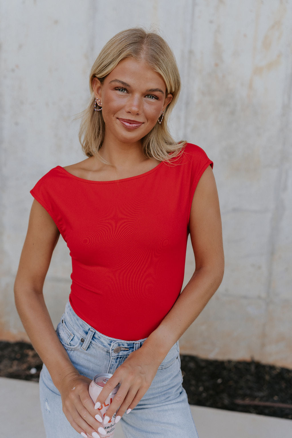 Upper body front view of female model wearing the Ryleigh Red Backless Bodysuit that has red knit fabric, an open back, and a scoop neckline. Worn with jeans.