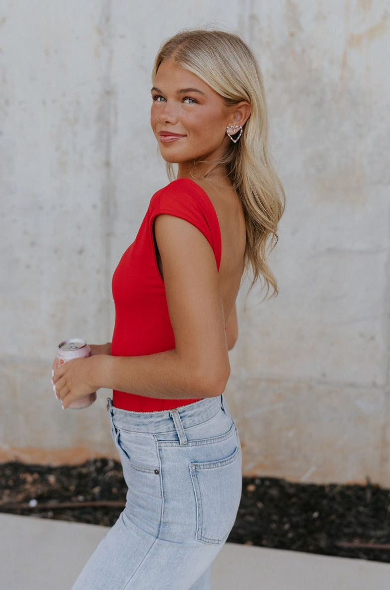 Upper body side view of female model wearing the Ryleigh Red Backless Bodysuit that has red knit fabric, an open back, and a scoop neckline. Worn with jeans.