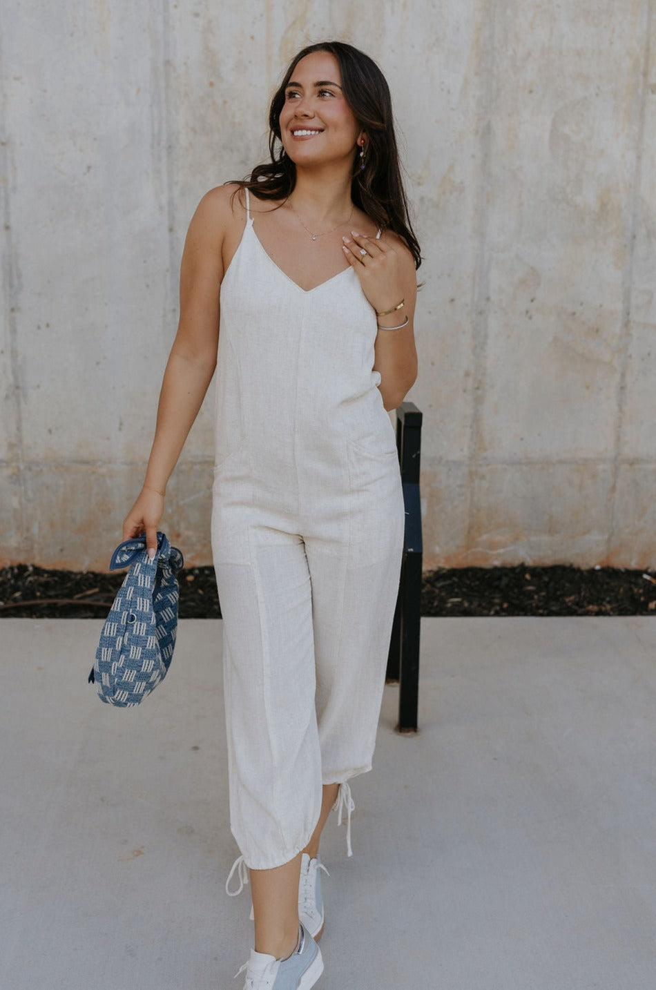 Full body front view of female model wearing the Parker Beige Linen Jogger Jumpsuit that has beige linen fabric, a v-neck, thin straps, pockets, and jogger style legs with ankle ties.