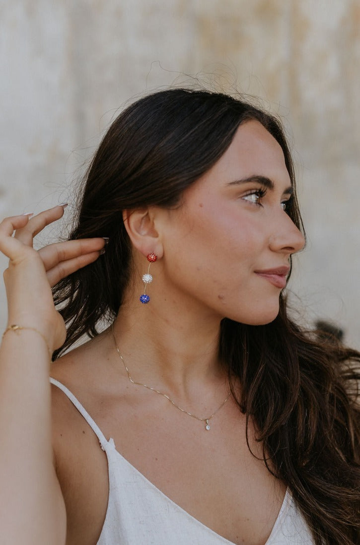 Side view of female model wearing the Paulina Red, White, & Blue Drop Earrings that have a red rhinestone sphere followed by white and blue spheres with gold wire between.