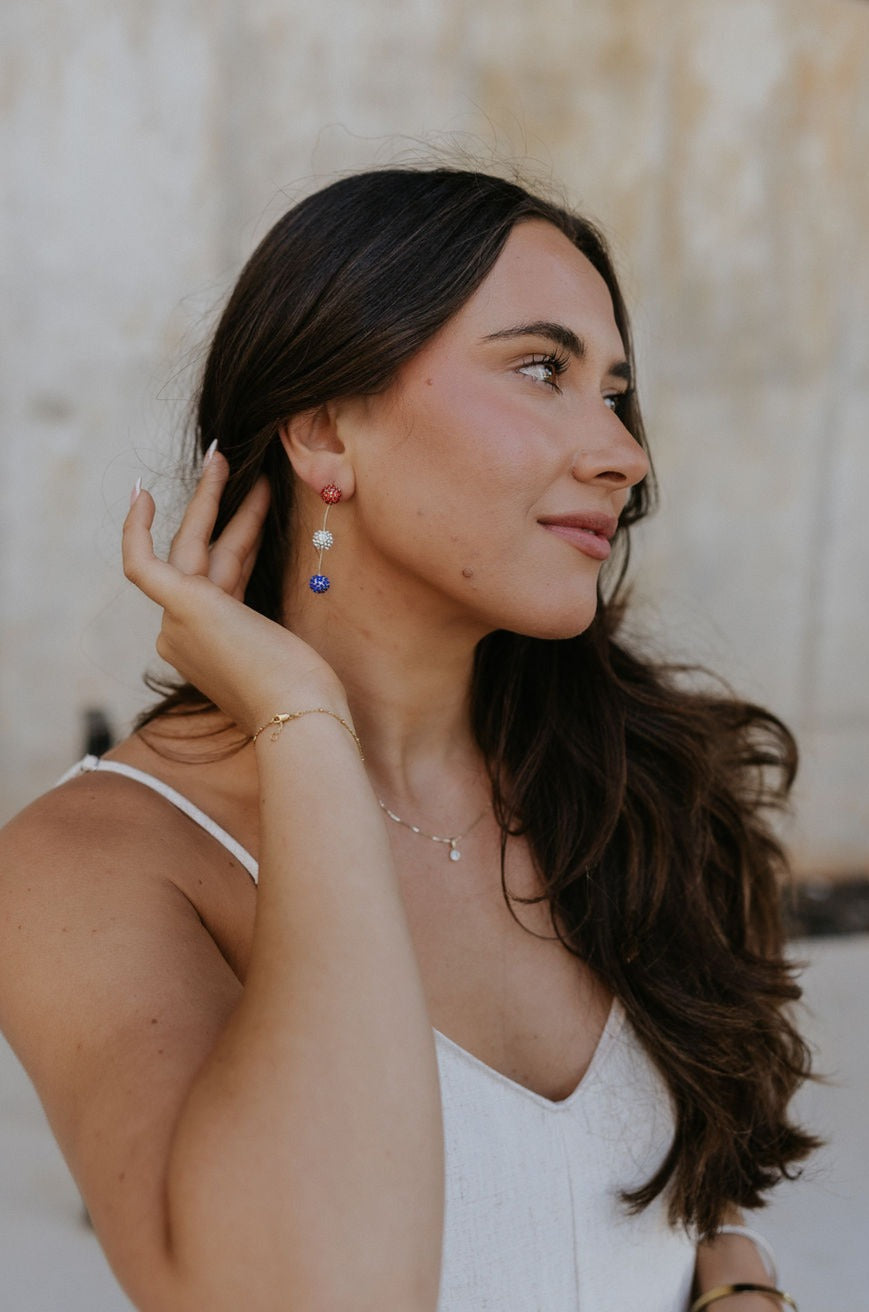 Side view of female model wearing the Paulina Red, White, & Blue Drop Earrings that have a red rhinestone sphere followed by white and blue spheres with gold wire between.