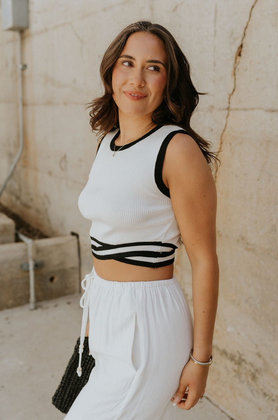 Side view of female model wearing the Lucia White & Black Groovy Hem Tank which features White Ribbed Fabric, Cropped Tank, Black Intertwine Hem Detail, Round Neckline and Sleeveless