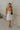 Full body back view of female model wearing the Tessa Natural Linen & Black Trim Mini Dress which features Natural Linen Fabric, Black Trim Details, Mini Length, Side Zipper, Halter Neckline with Tie Closure and Smocked Back
