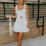 Front view of female model wearing the Destiny White Mini Dress.  The dress has a square neck with a ruffle detail and an adjustable belt with a ratan buckle.