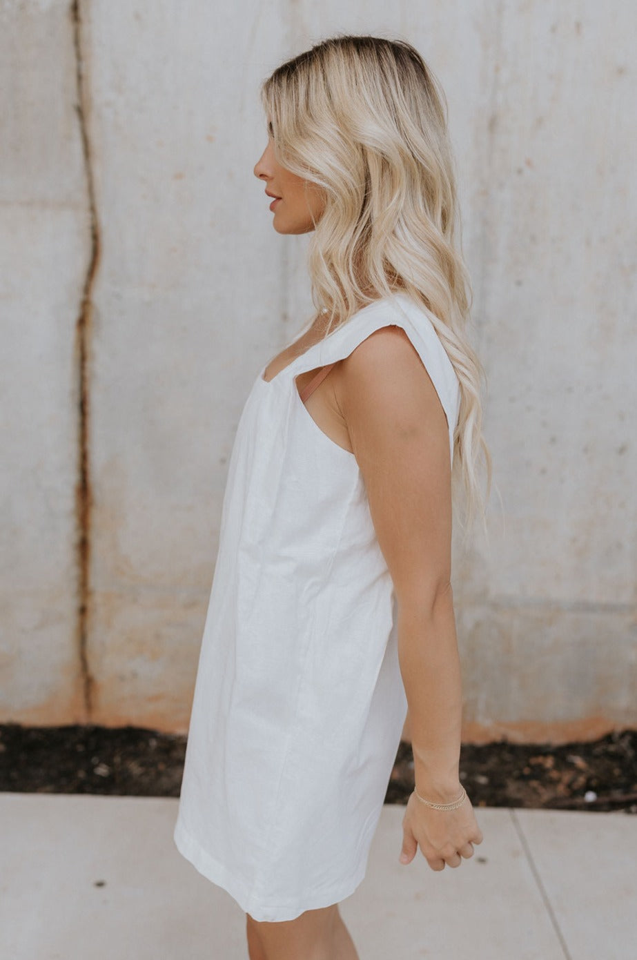Side view of female model wearing the Teagan White Square Neckline Romper which features White Linen Fabric, White Lining. Square Neckline, Sleeveless and Monochrome Back Zipper with Hook Closure