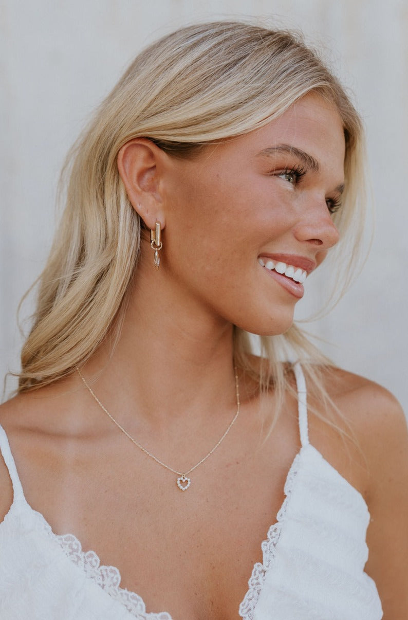 Model is shown turned to the side to reveal the Venus Earrings that have huggie frames with a heart pendant.