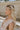 Side view of female model wearing the Emilia Satin Plunge Neckline Sleeveless Bodysuit which features Satin Fabric, Surplice Neckline with Button Snap, Sleeveless and Thong Bottom with Button Closure. the bodysuit is available in taupe and white