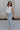 Full body side view of female model wearing the Emilia Satin Plunge Neckline Sleeveless Bodysuit which features Satin Fabric, Surplice Neckline with Button Snap, Sleeveless and Thong Bottom with Button Closure. the bodysuit is available in taupe and white