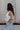 Side view of female model wearing the Emilia Satin Plunge Neckline Sleeveless Bodysuit which features Satin Fabric, Surplice Neckline with Button Snap, Sleeveless and Thong Bottom with Button Closure. the bodysuit is available in taupe and white