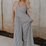 Full body view of female model wearing the Palmer Black & Cream Stripe Sleeveless Jumpsuit which features Black and Cream Linen Stripe Pattern, Wide Pant Legs, Side Pockets, Sweetheart Neckline,  Side Zipper, Adjustable Straps and Smocked Back