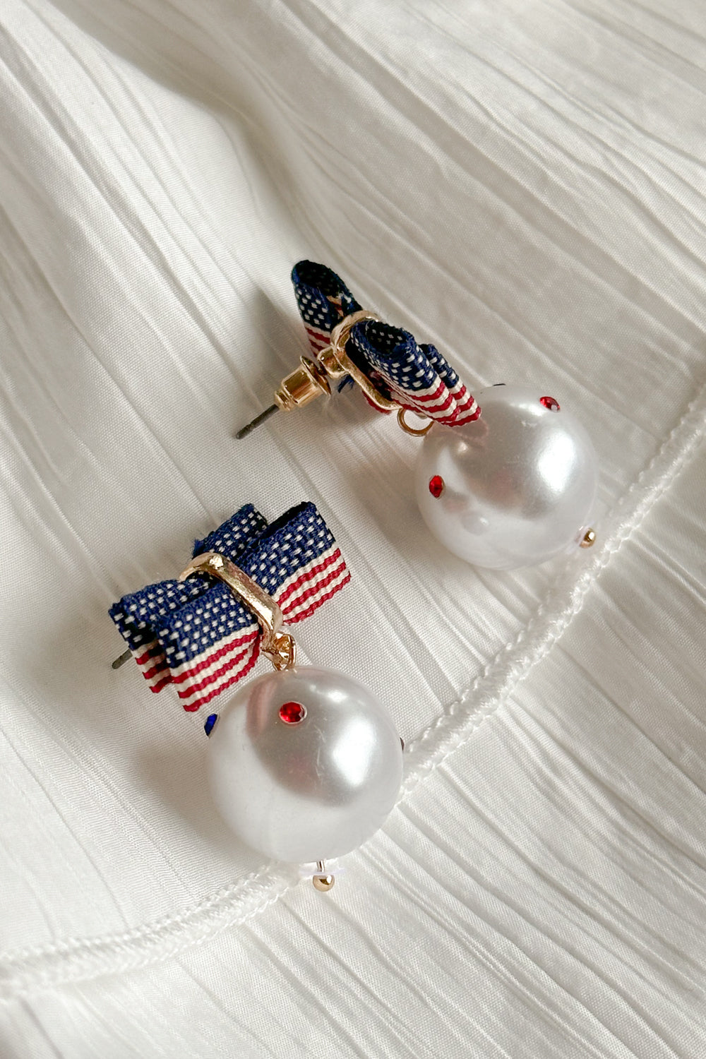 Close-up image of the Betsy Red, White, & Blue Pearl Earrings against a white background. Earrings have american flag bows and a pearl pendant with red white and blue stones. One is face up and one is sideways.