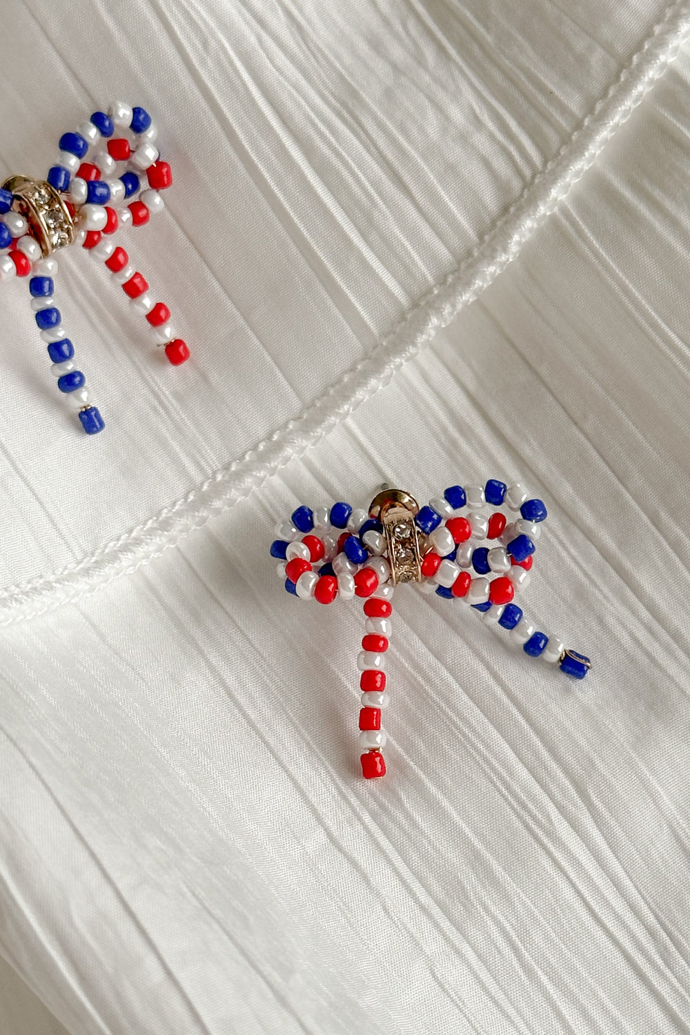 Image shows Quinn Red, White, & Blue Bow Earrings against a white background. Earrings have red white and blue beads in the shape of a bow, and the center is gold with rhinestones.