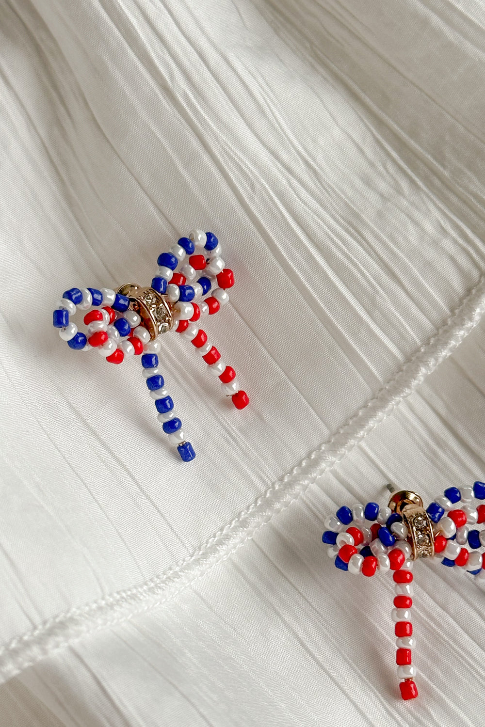 Image shows close-up of Quinn Red, White, & Blue Bow Earrings against a white background. Earrings have red white and blue beads in the shape of a bow, and the center is gold with rhinestones.