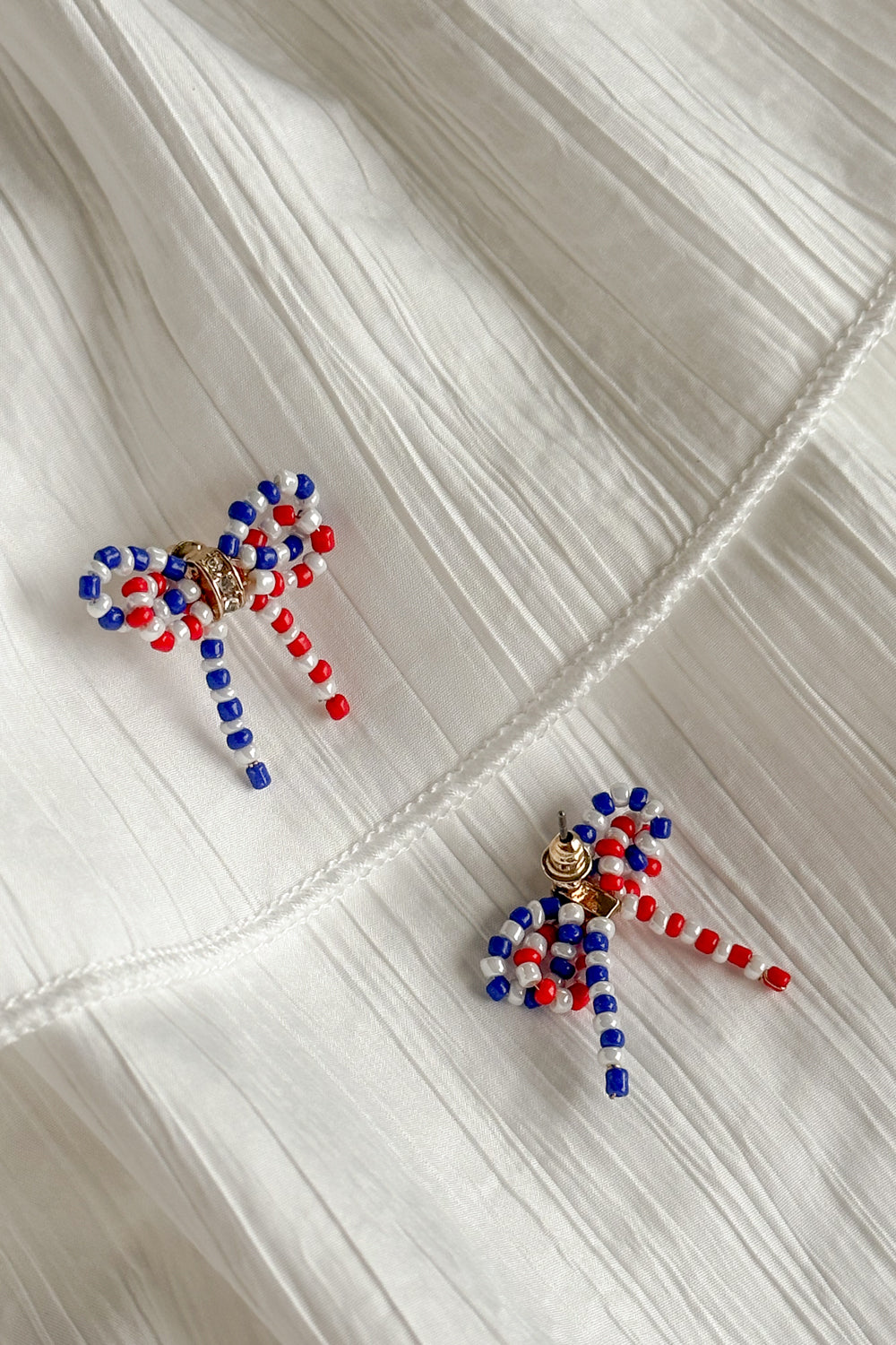 Image shows Quinn Red, White, & Blue Bow Earrings against a white background. Earrings have red white and blue beads in the shape of a bow, and the center is gold with rhinestones. 1 earring face down in image