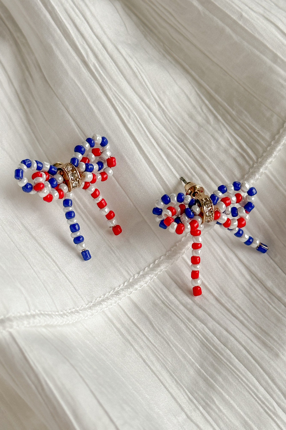 Image shows Quinn Red, White, & Blue Bow Earrings against a white background. Earrings have red white and blue beads in the shape of a bow, and the center is gold with rhinestones.
