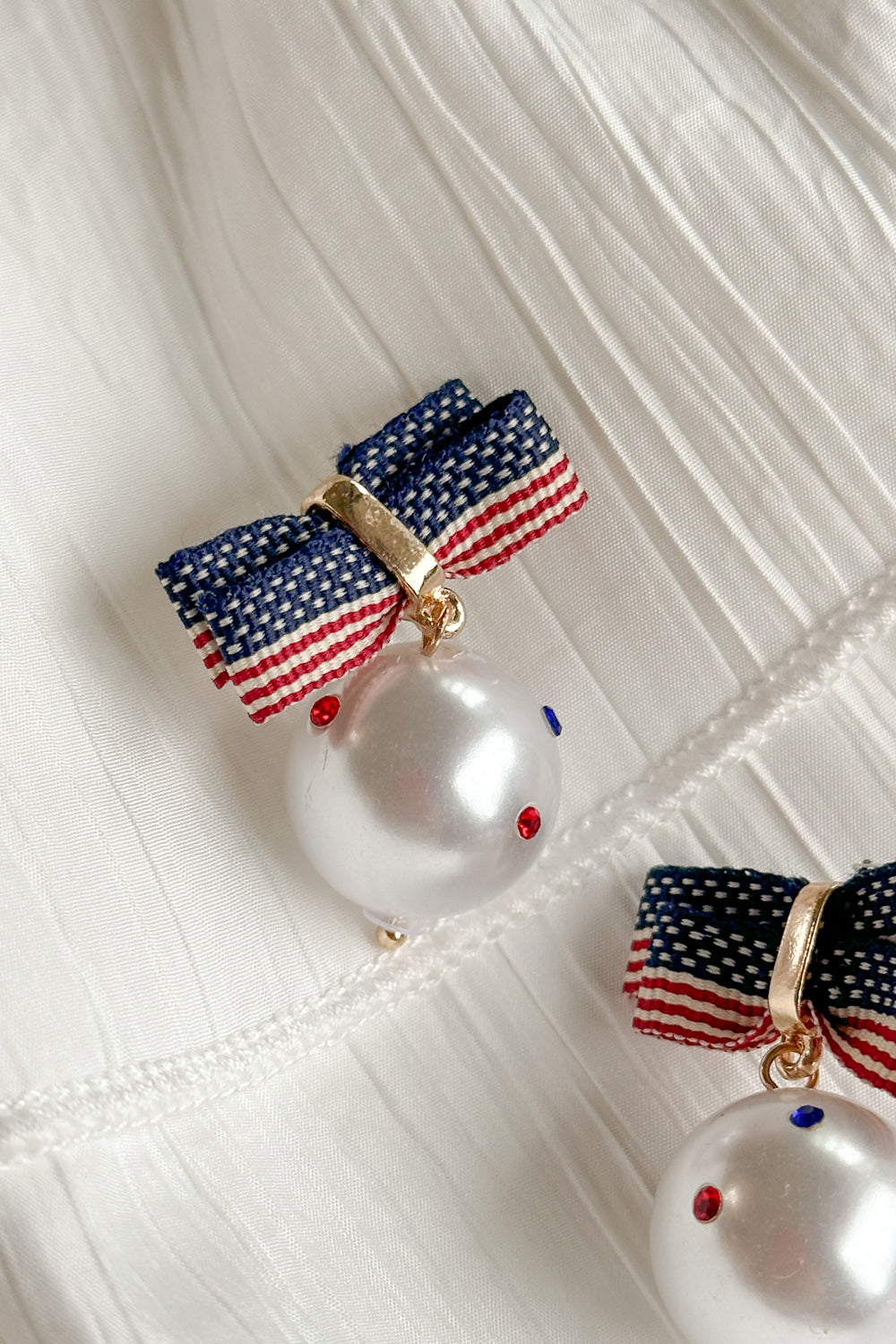 Close-up image of the Betsy Red, White, & Blue Pearl Earrings against a white background. Earrings have american flag bows and a pearl pendant with red white and blue stones.