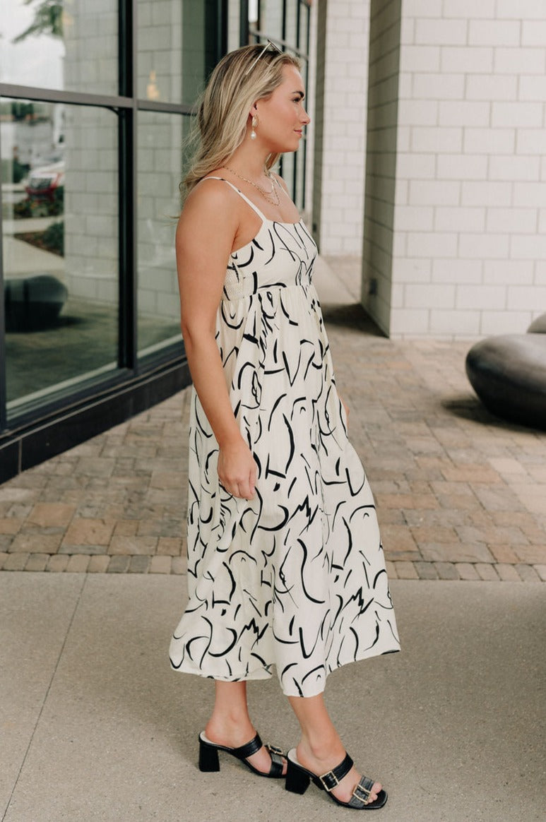 Full body side view of female model wearing the Luna Ivory & Black Abstract Midi Dress which features Cream and Black Lightweight Fabric, Abstract Print, Midi Length, Cream Lining, Square Neckline, Adjustable Straps and Smocked Back