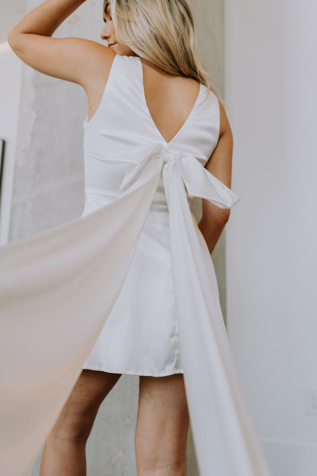 Back view of female model wearing the Melissa White Satin Bow Mini Dress which features White Satin Fabric, White Lining, Mini Length, Round Neckline, Sleeveless, Side Monochrome Zipper with Hook Closure and Bow Detail