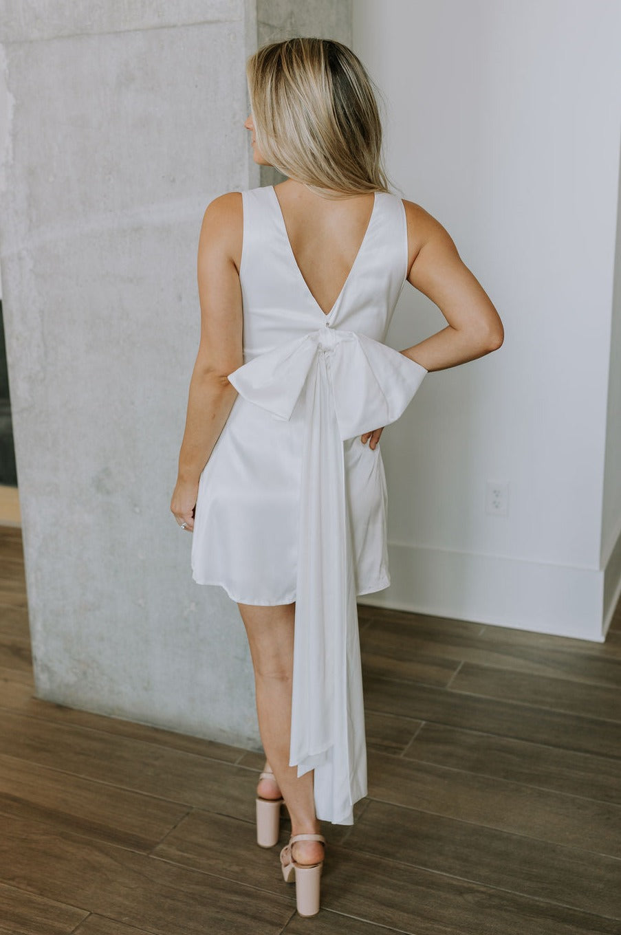 back view of female model wearing the Melissa White Satin Bow Mini Dress which features White Satin Fabric, White Lining, Mini Length, Round Neckline, Sleeveless, Side Monochrome Zipper with Hook Closure and Bow Detail