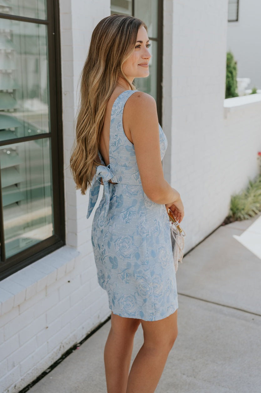side view of female model wearing the Amalia Light Blue Rose Bow Mini Dress which features Blue and White Rose Floral Print, Blue Lining, Mini Length, Plunge Neckline, Sleeveless, Back Bow Detail, Back Cutout and Monochrome Back Zipper with Hook Closure