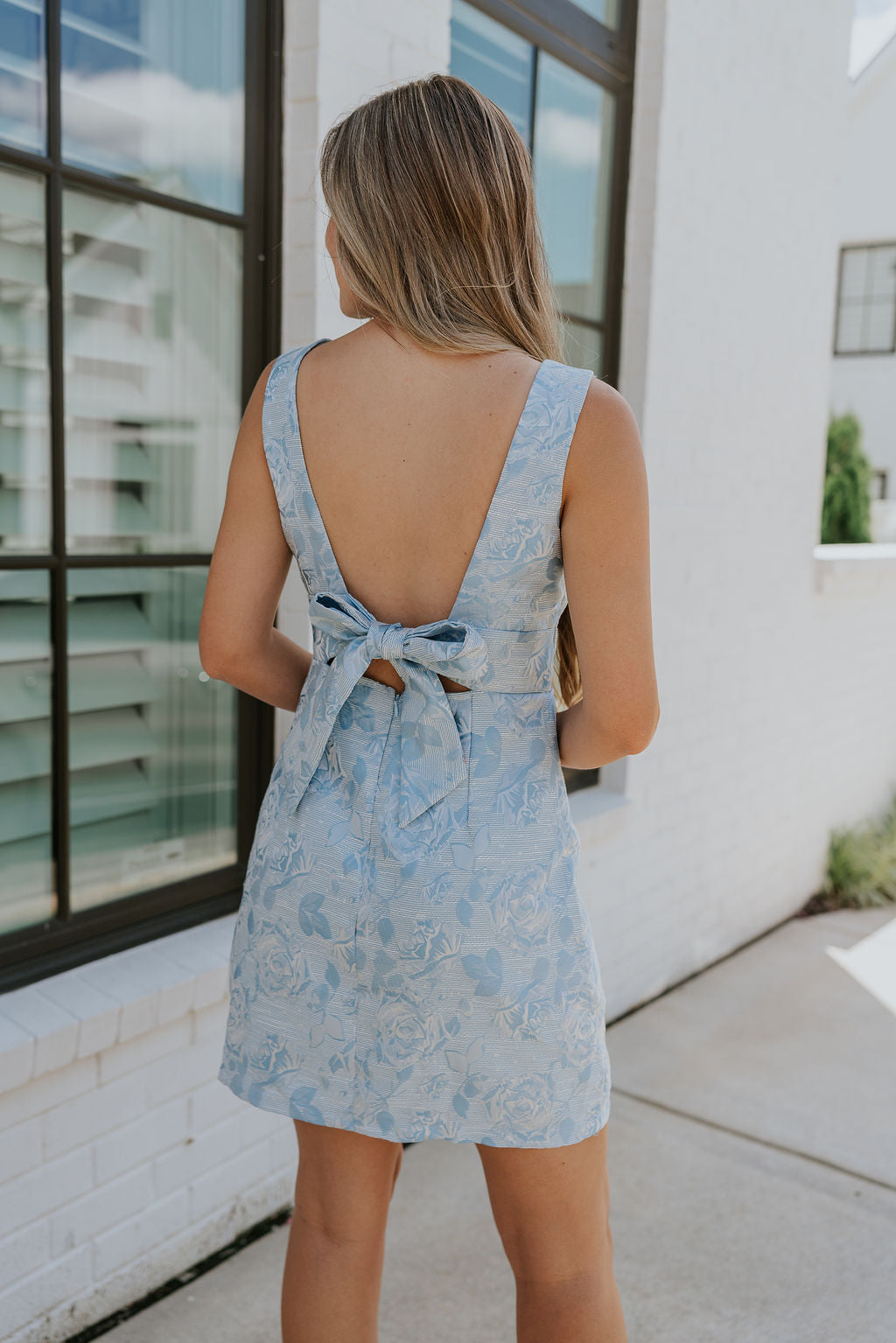 back view of female model wearing the Amalia Light Blue Rose Bow Mini Dress which features Blue and White Rose Floral Print, Blue Lining, Mini Length, Plunge Neckline, Sleeveless, Back Bow Detail, Back Cutout and Monochrome Back Zipper with Hook Closure