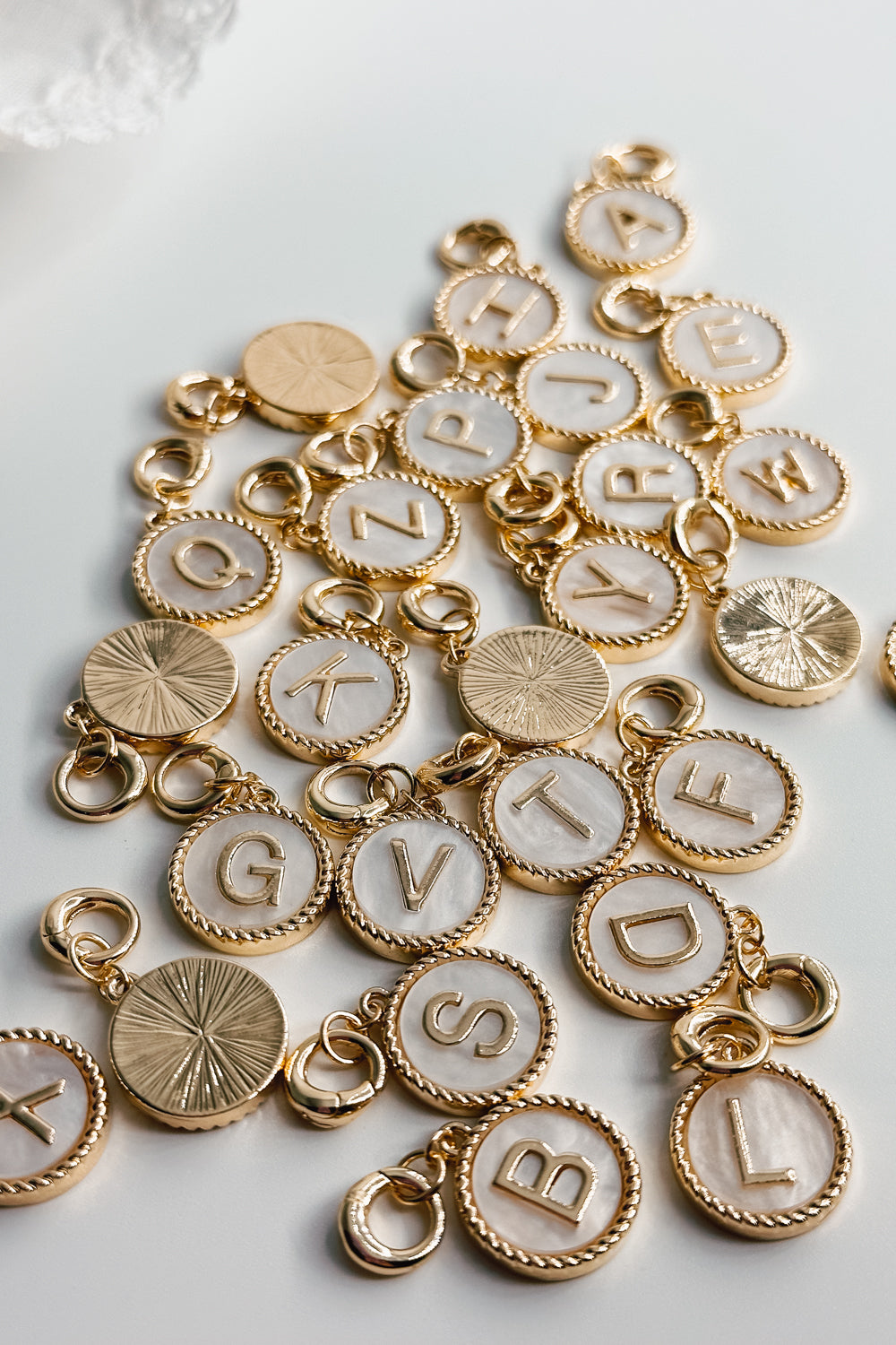 Image shows an assortment of the Alphabet Charms against a white background. Some charms are face up and some are face down. Charms are gold circles with white center and gold letter.