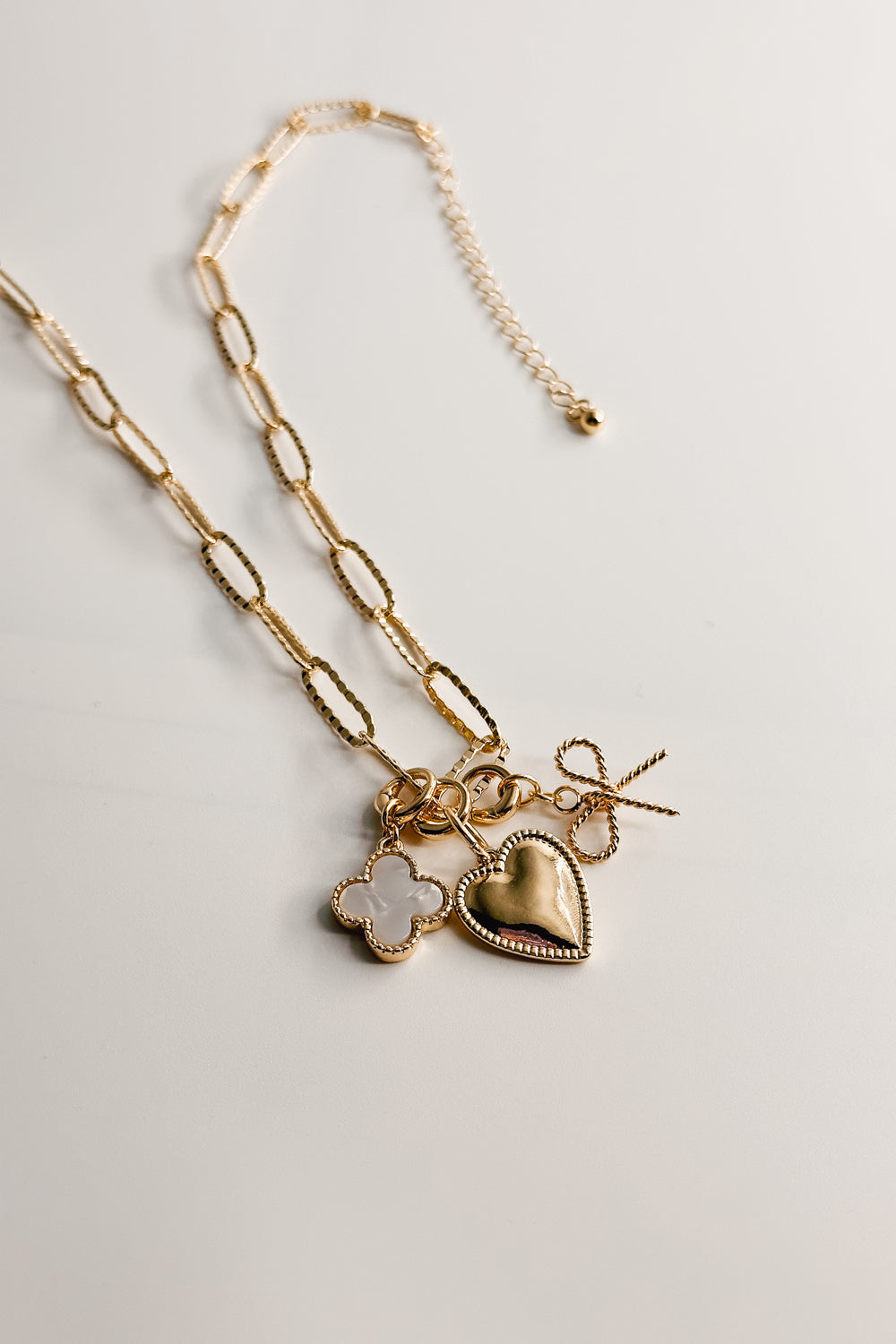 Image shows gold necklace chain with white clover charm, heart charm, and rope bow charm. Shown against a white background.