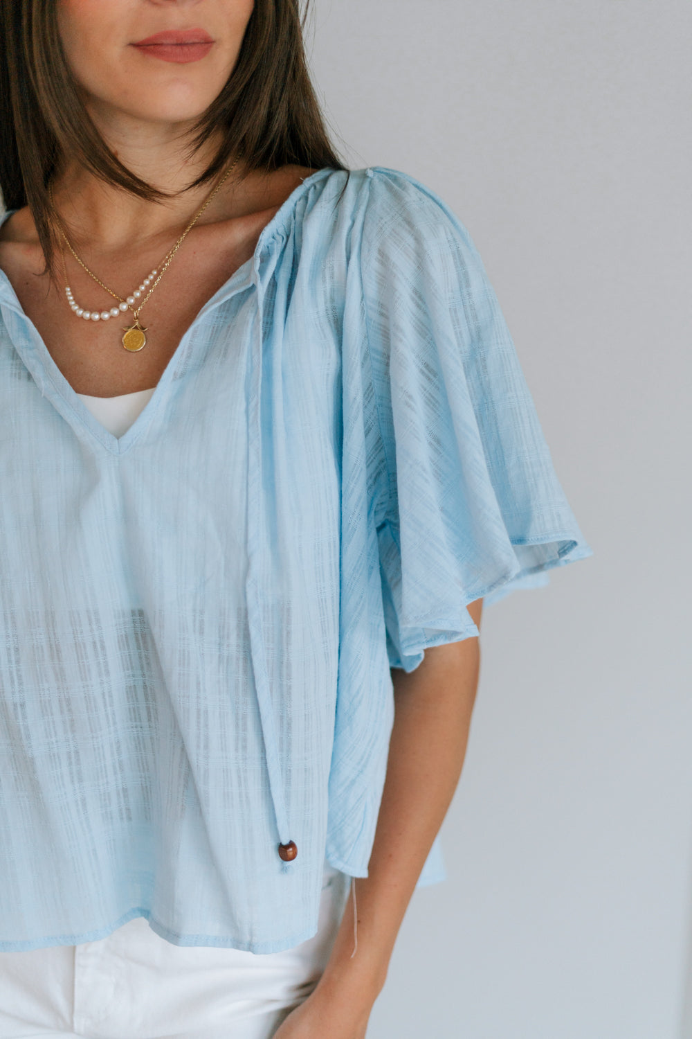 Close up view of female model wearing the Danna Light Blue Short Sleeve Top which features Light Blue Cotton Fabric, Round Neckline with Tie Closure, Wooden Beads Detail and Short Sleeves