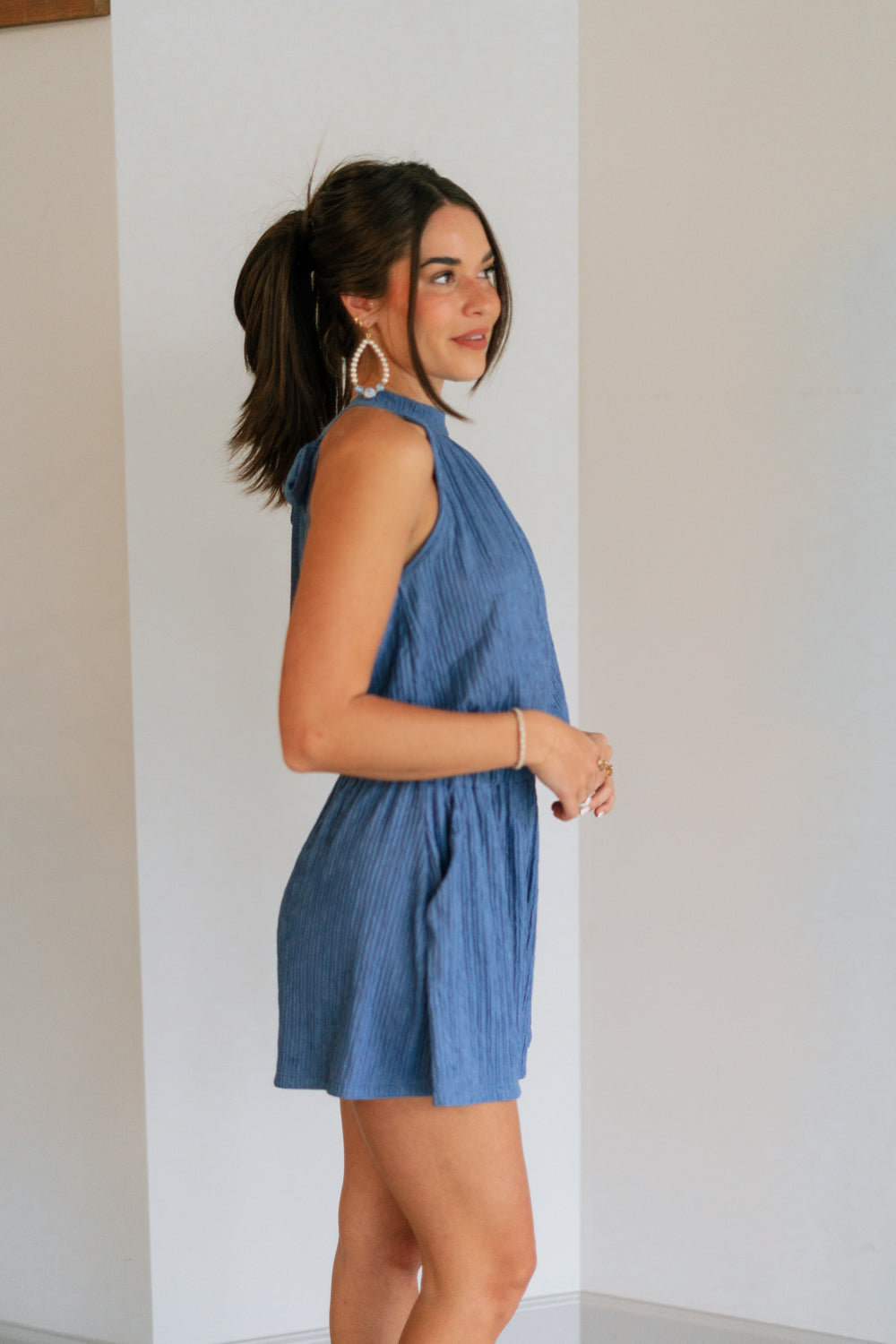 Side view of female model wearing the Leila Dark Blue Halter Neckline Romper which features Grey Blue Textured Fabric, Two Side Slit Pockets, Halter Neckline with Tie, Elastic Waistband and Back Key Hole