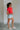 Full body side view of female model wearing the Evie Red Coral Short Sleeve Top which features Coral Red Cotton Fabric, Cropped Waist with Raw Hem, Round Neckline and Short Sleeves