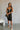 Full body front view of model wearing the Gianna Black & White Midi Dress that has black fabric, and white straps with white neckline bow trim. Model is wearing white heels and black purse.