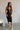Full body front view of model wearing the Gianna Black & White Midi Dress that has black fabric, and white straps with white neckline bow trim. Model is wearing white heels