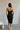 Full body back view of model wearing the Gianna Black & White Midi Dress that has black fabric, and white straps with white neckline bow trim. Model is wearing white heels