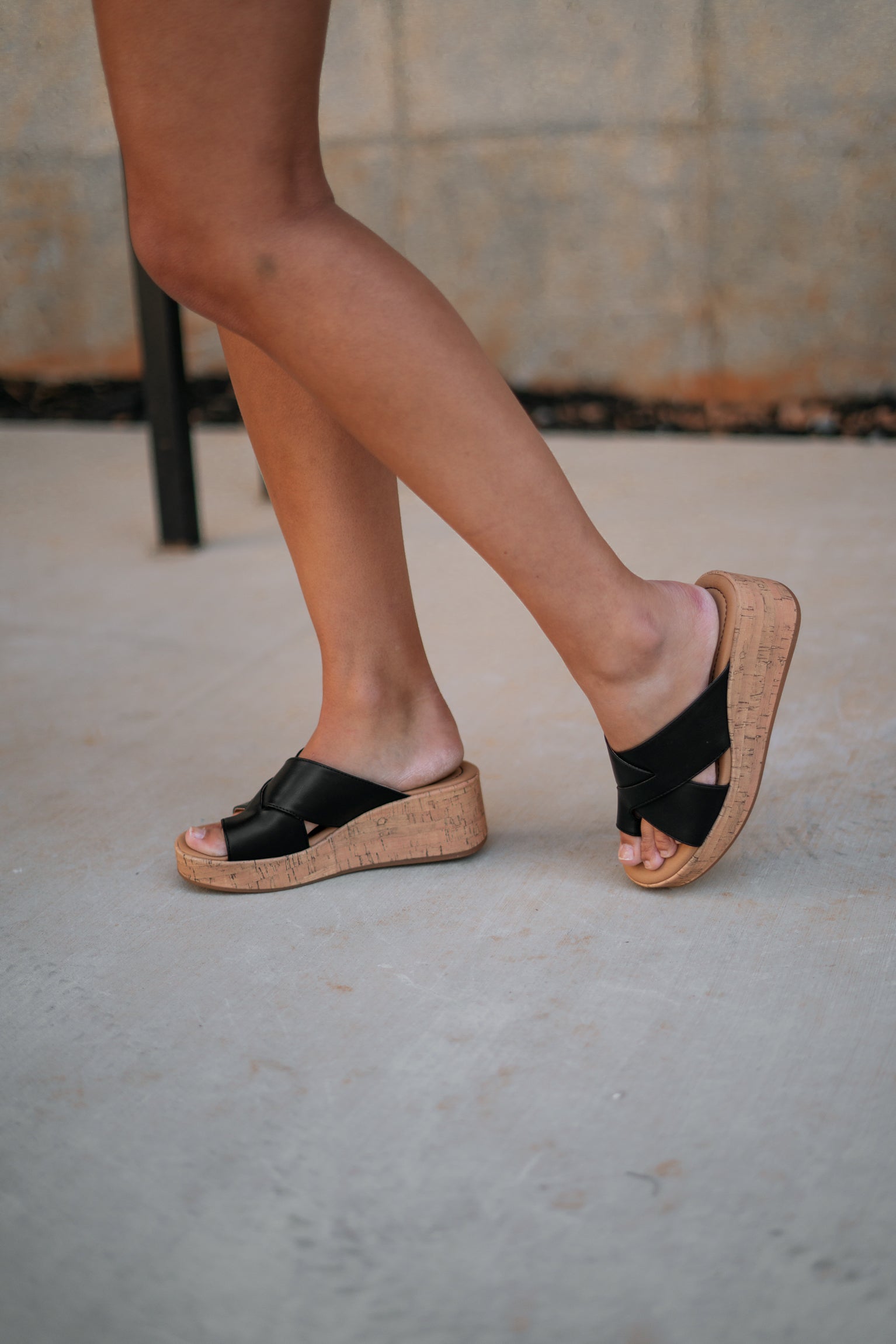 Right side view of female model wearing the Sunny Wedge Sandal in Black which features Black Faux Leather Upper, Slip-On Style and Criss-Cross Strap & Toe Wrap Detail