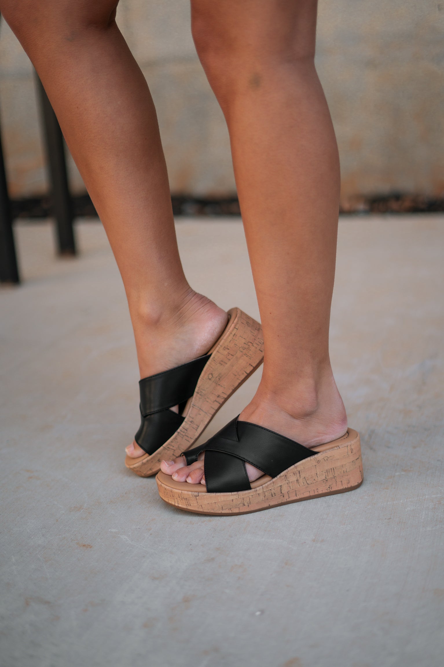 Right side view of female model wearing the Sunny Wedge Sandal in Black which features Black Faux Leather Upper, Slip-On Style and Criss-Cross Strap & Toe Wrap Detail