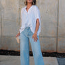 Full body front view of female model wearing the Thea White Lightweight Top that has lightweight white fabric with subtle monochrome stripes, oversized fit, v neckline, and short batwing sleeves. Shirt is front tucked into jeans.