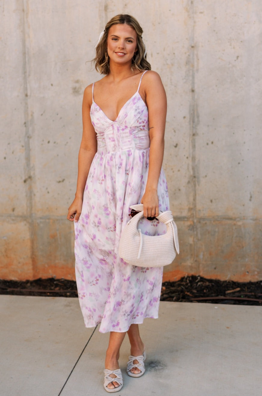 Full body front view of female model wearing the Bridget Lavender Watercolor Midi Dress that has a lavender watercolor floral pattern, a corset top, thin straps, and midi length