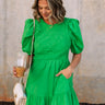 front view of female model wearing the Lea Green Puff Sleeve Mini Dress that has kelly green fabric, a smocked upper with high neck, short puff sleeves, and mini length. Model is holding coffee and has purse on shoulder.
