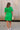 Full body back view of female model wearing the Lea Green Puff Sleeve Mini Dress that has kelly green fabric, a smocked upper with high neck, short puff sleeves, and mini length. Model is holding coffee and has purse on shoulder.