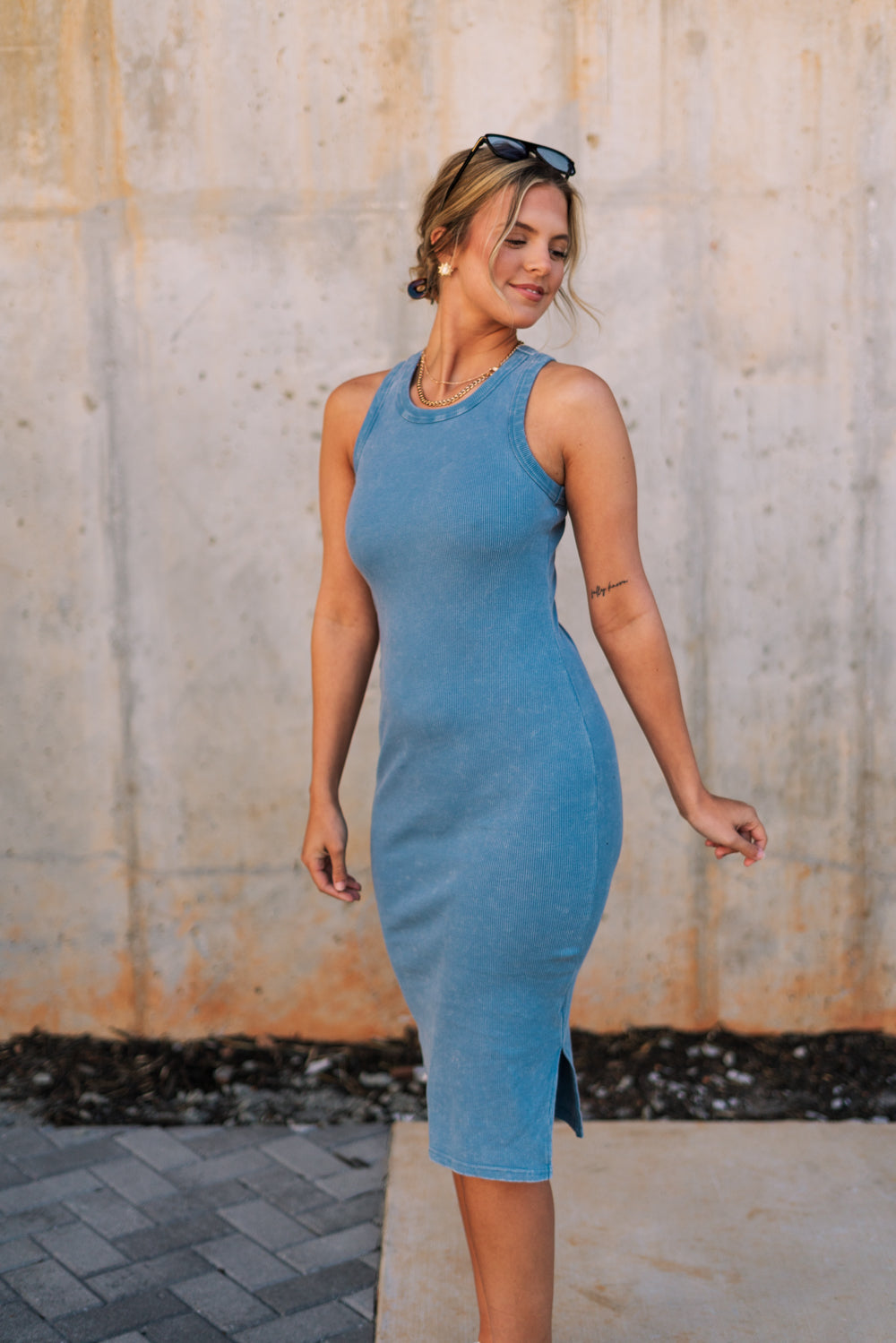 Side view of female model wearing the Paula Blue Ribbed Midi Dress that has ribbed medium blue fabric, a round neck, sleeveless, and midi length hem. 