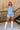 Full body front view of female model wearing the Georgia Belted Denim Romper in light wash, that has a button up front, collar, and belted waist. Paired with white boots.