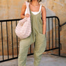 Full body front view of female model wearing the Alicia Olive Knit Jumpsuit that has olive green knit fabric, large front pockets, tapered legs, and thin straps. Styled over a white tank top.