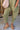 Lower body front view of female model wearing the Alicia Olive Knit Jumpsuit that has olive green knit fabric, large front pockets, tapered legs, and thin straps. Styled over a white tank top.