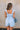 Back view of female model wearing the Taylor Light Wash Denim Romper that has light wash denim fabric, straps with frayed hems, elastic waist, neckline pleating, and side pockets.