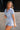 Full body side view of female model wearing the Mavis Denim Short Sleeve Zip-Up Romper which features Denim Fabric, Two Front Pockets, Front Zipper Closure, Adjustable Monochrome Belt with Loops, Collared Neckline and Short Sleeves