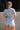 Back view of female model wearing the Mavis Denim Short Sleeve Zip-Up Romper which features Denim Fabric, Two Front Pockets, Front Zipper Closure, Adjustable Monochrome Belt with Loops, Collared Neckline and Short Sleeves