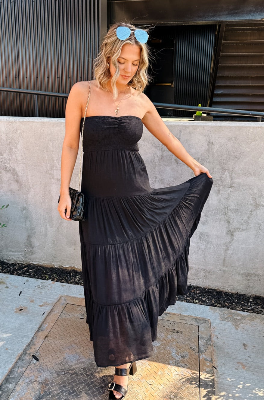 Full body front view of female model wearing the Amaya Black Strapless Maxi Dress that has a smocked upper and a tiered skirt. Model is holding skirt
