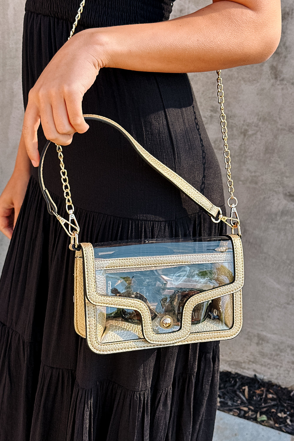 Close up side view of female model wearing the Miley Clear Purse in Gold which features clear with leather details, gold snap closure, and an adjustable gold chain strap with gold details.