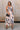 Full body front view of female model wearing the Elina Printed Button Front Midi Dress that has ivory, black, and orange geometric printing, a tie belt, a button up front, a collared sleeveless neckline, and midi length.