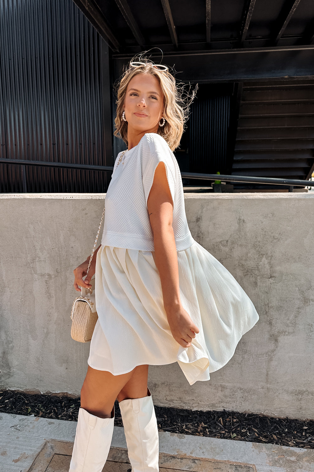 Side view of female model wearing the Kara Cream & White Twofer Mini Dress that has a texture white upper with short sleeves and a cream skirt with pleating. Worn with white boots.